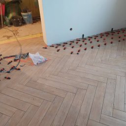 FachGro usługi remontowe - Układanie parkietu w jodełkę z użyciem klipsów poziomujących. Widoczne narzędzia i materiały budowlane. Jasne płytki imitujące drewno. Częściowo wykończone pomieszczenie.