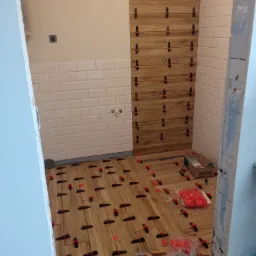 Łazienka w trakcie remontu: układanie drewnopodobnych płytek na podłodze i ścianie, widoczne krzyżyki dystansowe, poziomica i narzędzia glazurnicze.