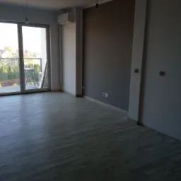 Pomalowane na szaro ściany i ułożona podłoga z paneli imitujących drewno w pustym pokoju z dużym oknem balkonowym w Wolicy.