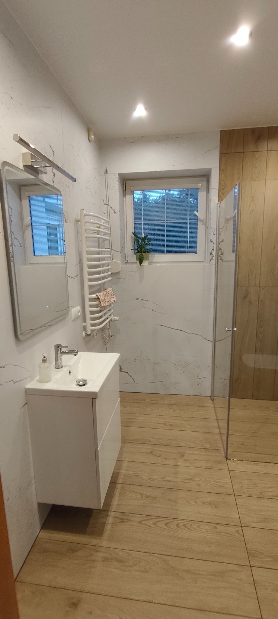 Jasna łazienka z drewnopodobną podłogą i marmurowymi ścianami. Umywalka z szafką, grzejnik drabinkowy i kabina prysznicowa z przezroczystego szkła.