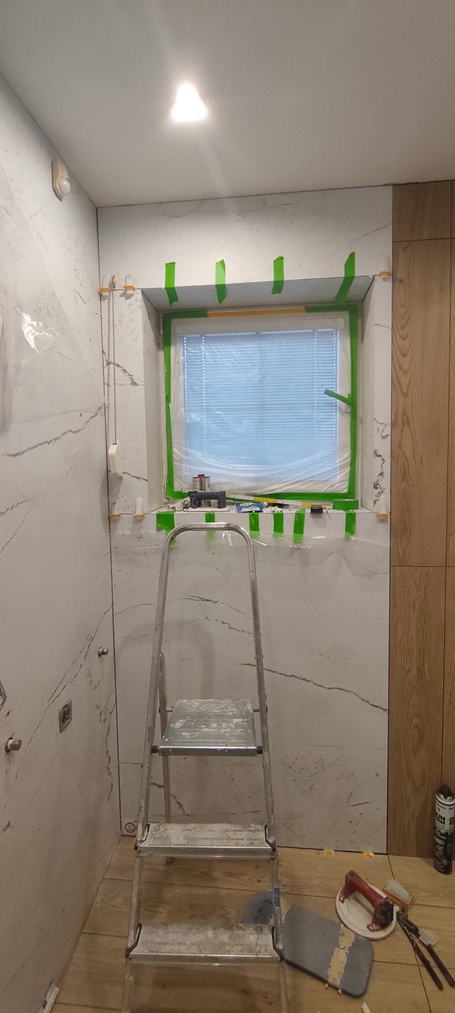 Łazienka w trakcie wykańczania: marmurowe płytki na ścianach, okno zabezpieczone taśmą, drabina i narzędzia glazurnicze na podłodze z paneli.