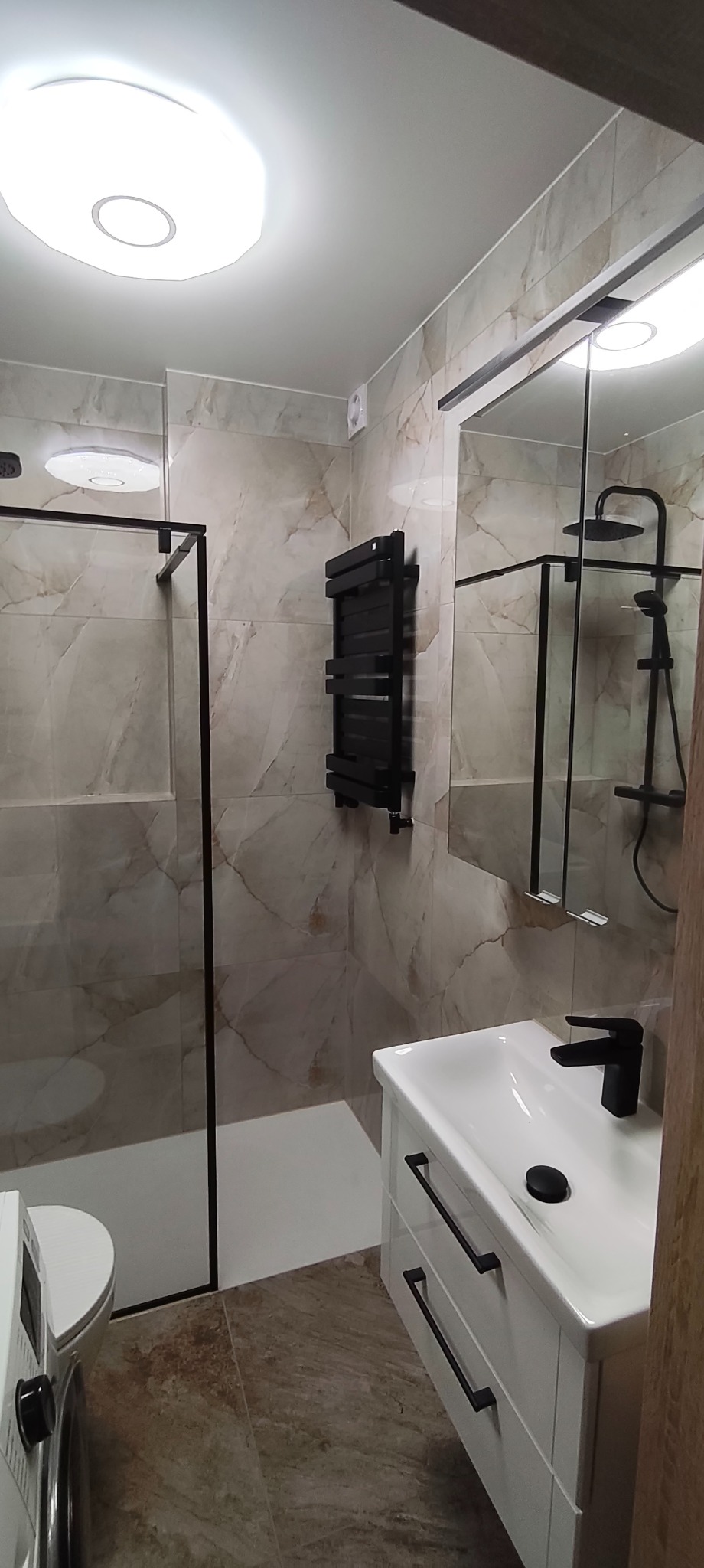 Wykończona łazienka z kabiną prysznicową typu walk-in, umywalką z szafką i grzejnikiem drabinkowym w czarnym kolorze. Płytki imitujące marmur na ścianach i podłodze.