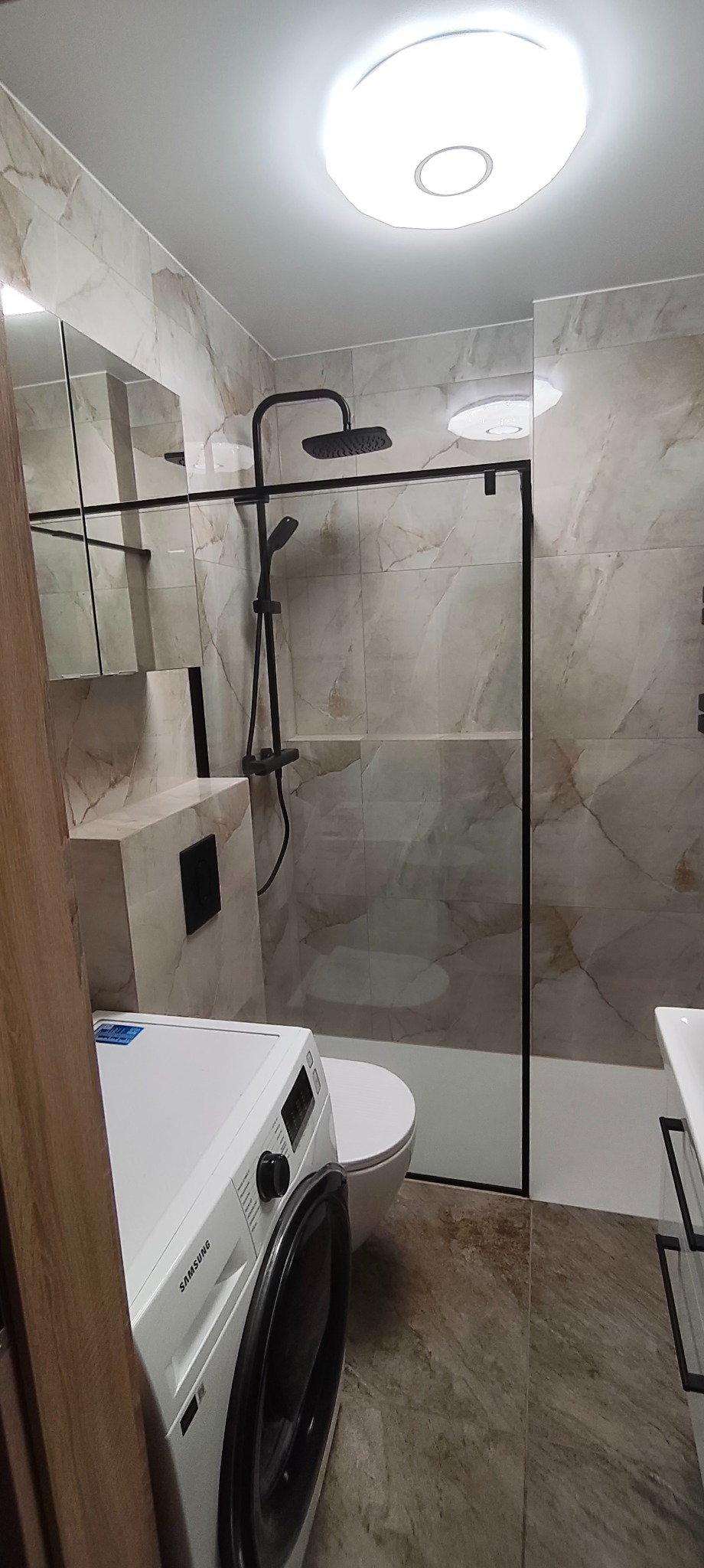 Wykończona łazienka z kabiną typu walk-in, płytkami imitującymi marmur, czarną armaturą i pralką Samsung. Nowoczesny design, oświetlenie sufitowe.