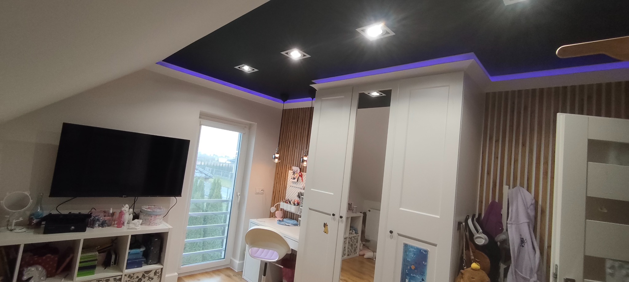 Pokój na poddaszu z czarnym sufitem, oświetleniem LED i drewnianą ścianą. Widoczne biurko, szafa z lustrem i telewizor. Nowoczesny design z elementami dekoracyjnymi.