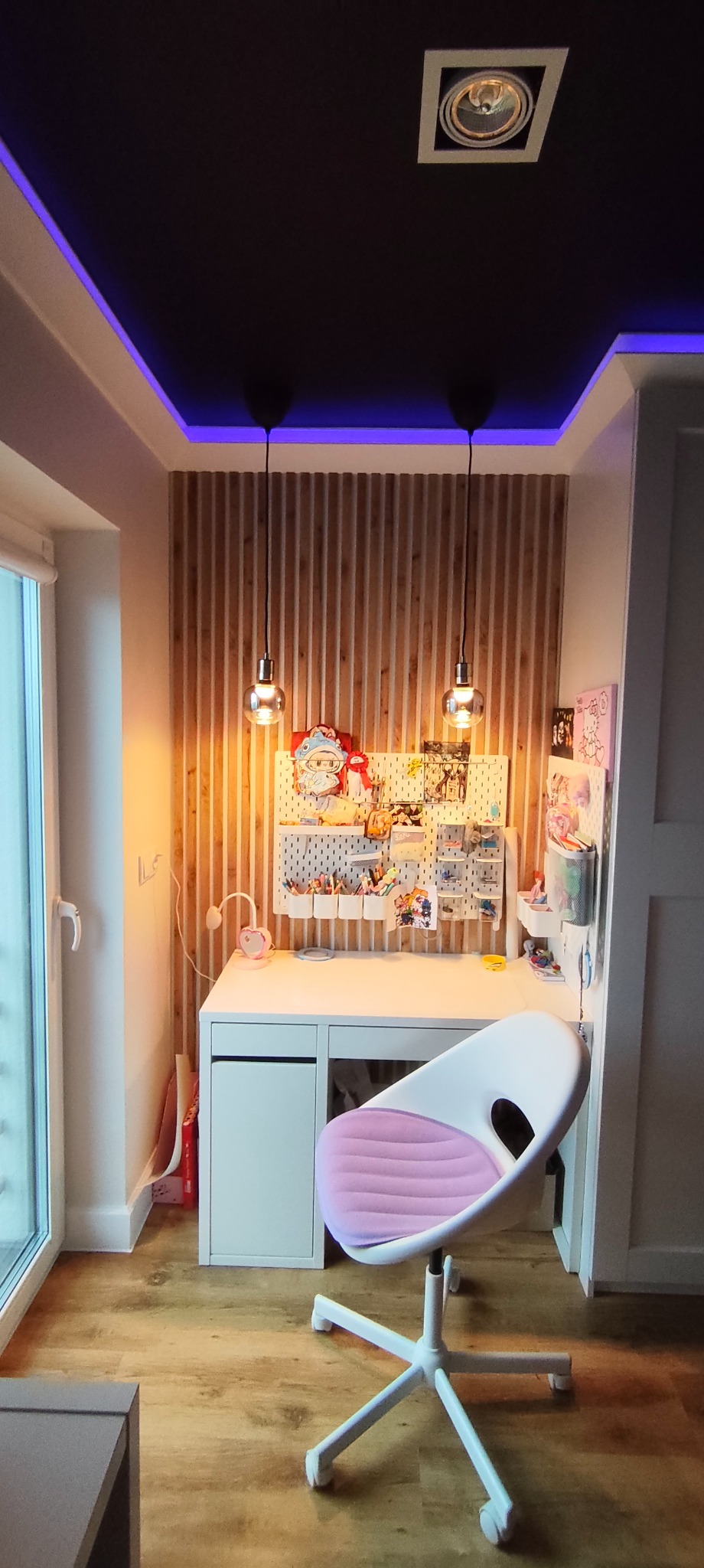 Pokój młodzieżowy z biurkiem, drewnianą ścianą z lamelami i podwieszanym sufitem z niebieskim oświetleniem LED. Na biurku tablica z akcesoriami.