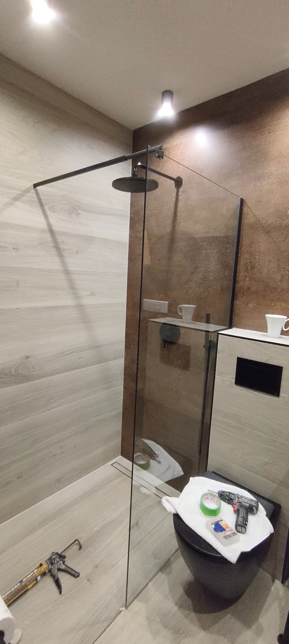 Nowoczesna kabina prysznicowa typu walk-in z czarną armaturą i szklaną ścianką. Na sedesie leżą narzędzia i akcesoria montażowe. Widoczne płytki imitujące drewno.
