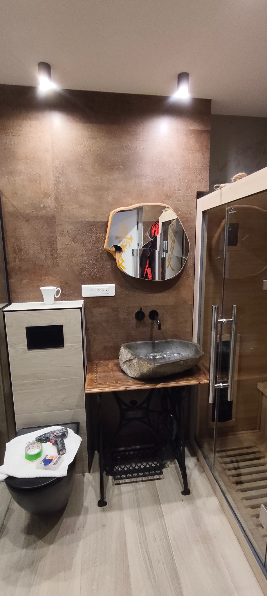 Nowoczesna łazienka z umywalką z kamienia na podstawie maszyny do szycia Singer, lustro w drewnianej ramie o nieregularnym kształcie, brązowe płytki na ścianach.