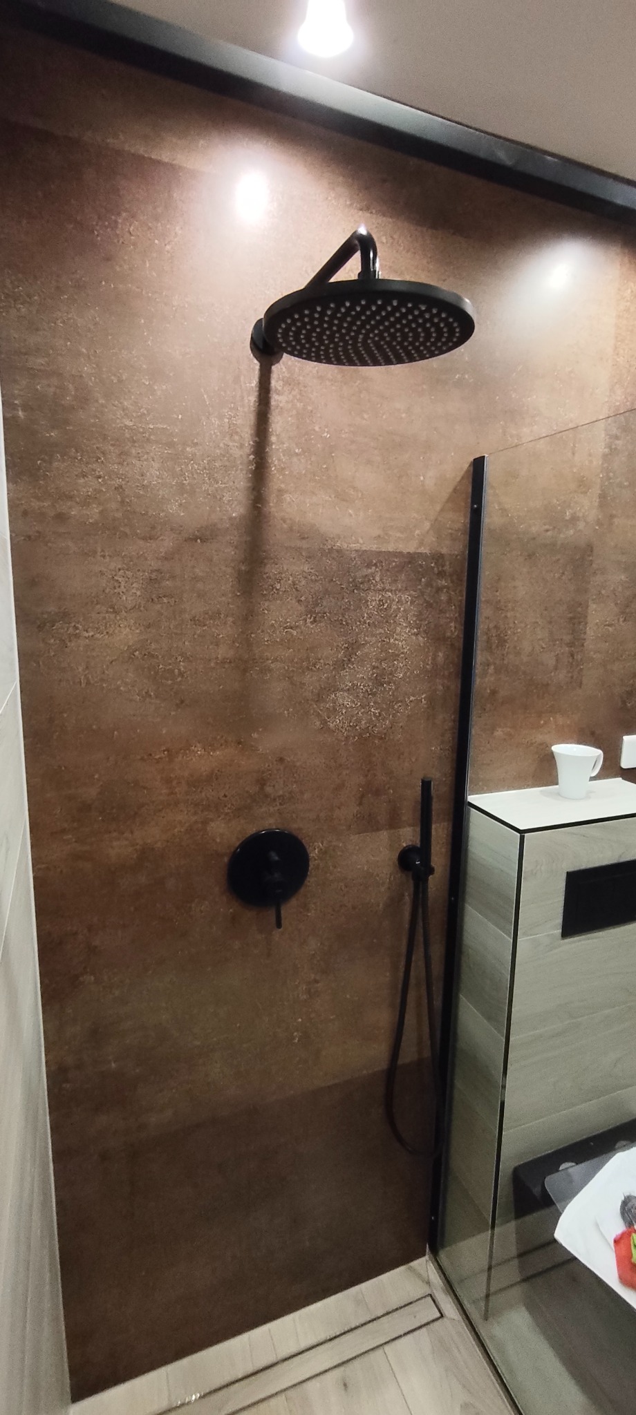 Nowoczesna kabina prysznicowa walk-in z brązowymi płytkami imitującymi kamień, czarną armaturą i szklaną ścianką. Widoczny odpływ liniowy i fragment szafki umywalkowej.