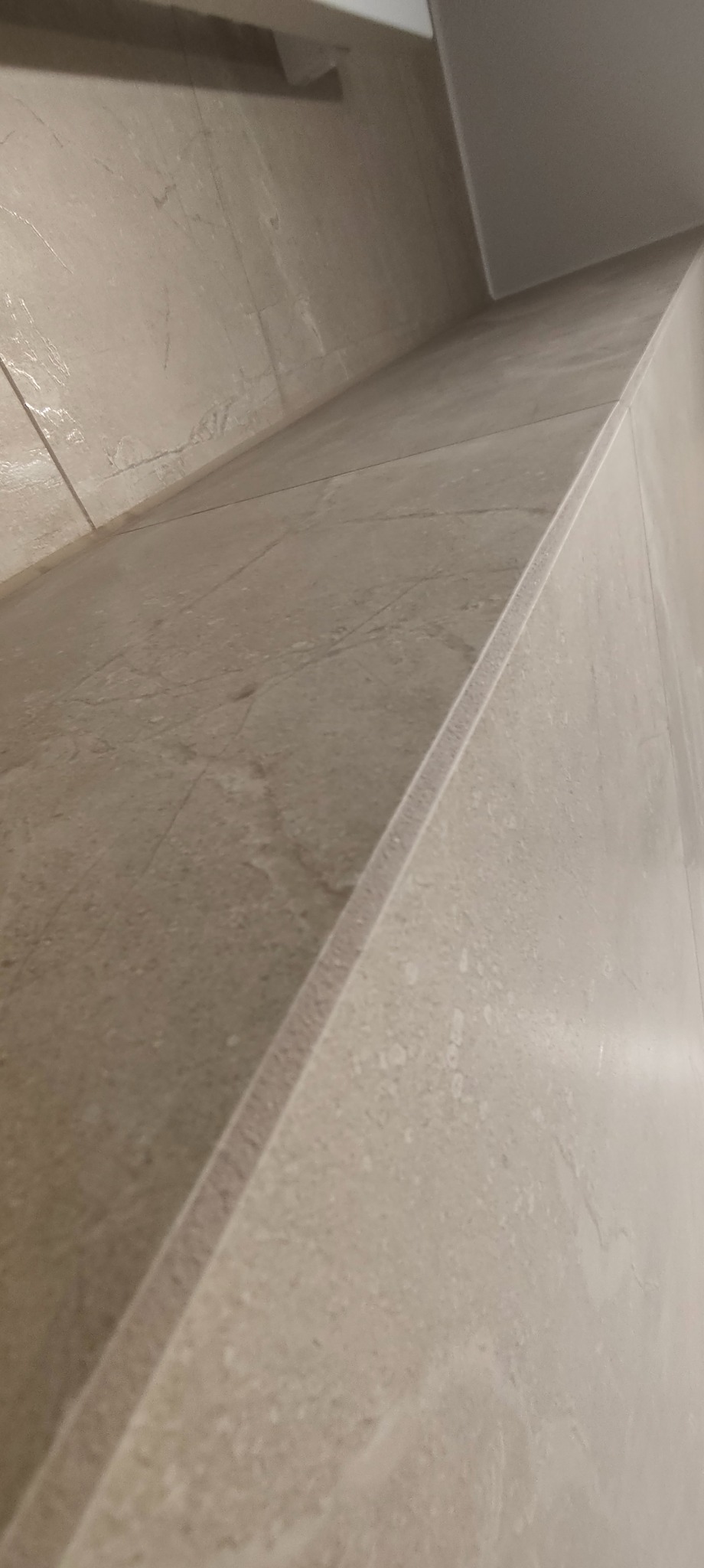 Beżowa, marmurkowa płytka ścienna w łazience, ułożona z precyzją, z widoczną krawędzią i fugą. Minimalistyczny design, ciepłe oświetlenie.