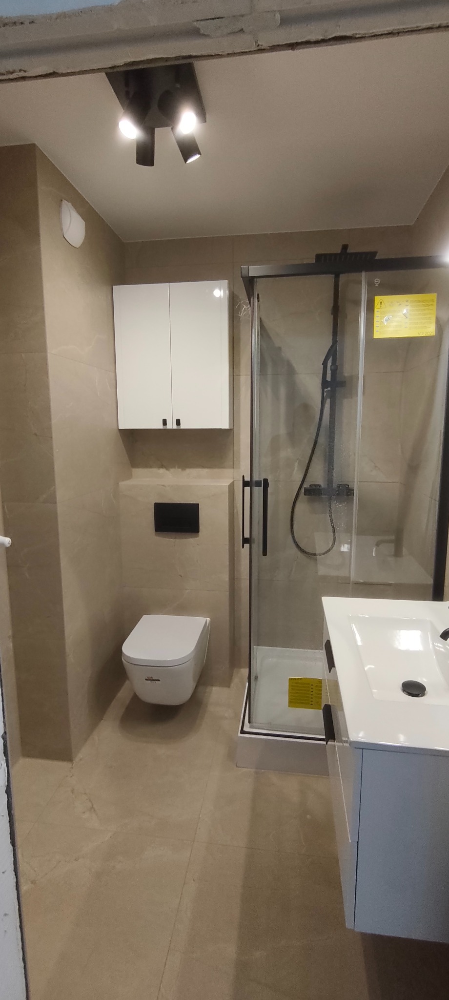 Nowoczesna łazienka z beżowymi płytkami, kabiną prysznicową z czarnymi ramami, podwieszaną toaletą i białą szafką z umywalką. Minimalistyczne oświetlenie.