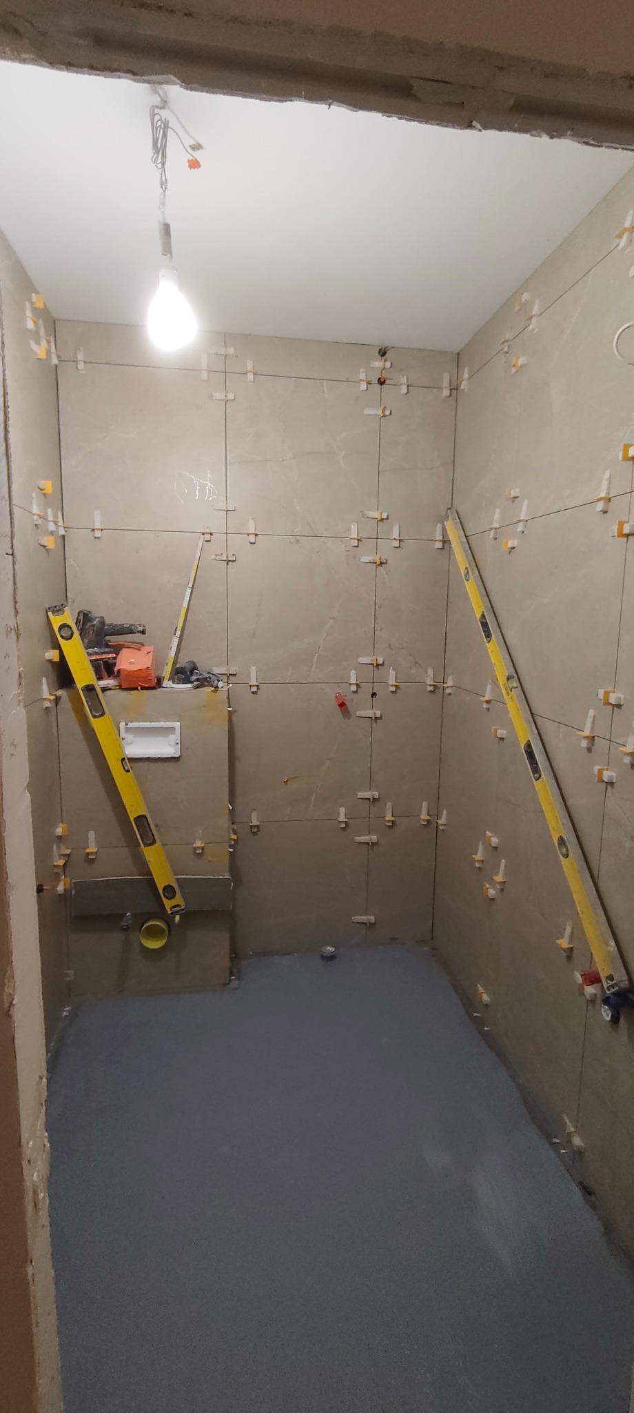 Wnętrze łazienki w trakcie prac glazurniczych: ułożone płytki na ścianach z użyciem krzyżyków dystansowych, poziomice, narzędzia i wylewka na podłodze.