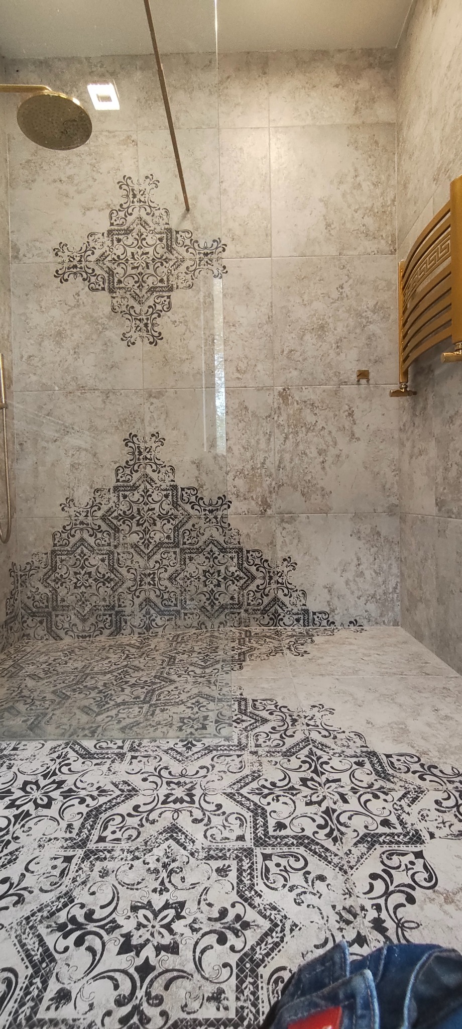 Wykończona kabina prysznicowa z beżowymi płytkami zdobionymi czarnym, orientalnym wzorem. Złoty grzejnik drabinkowy i deszczownica. Nowoczesny design.