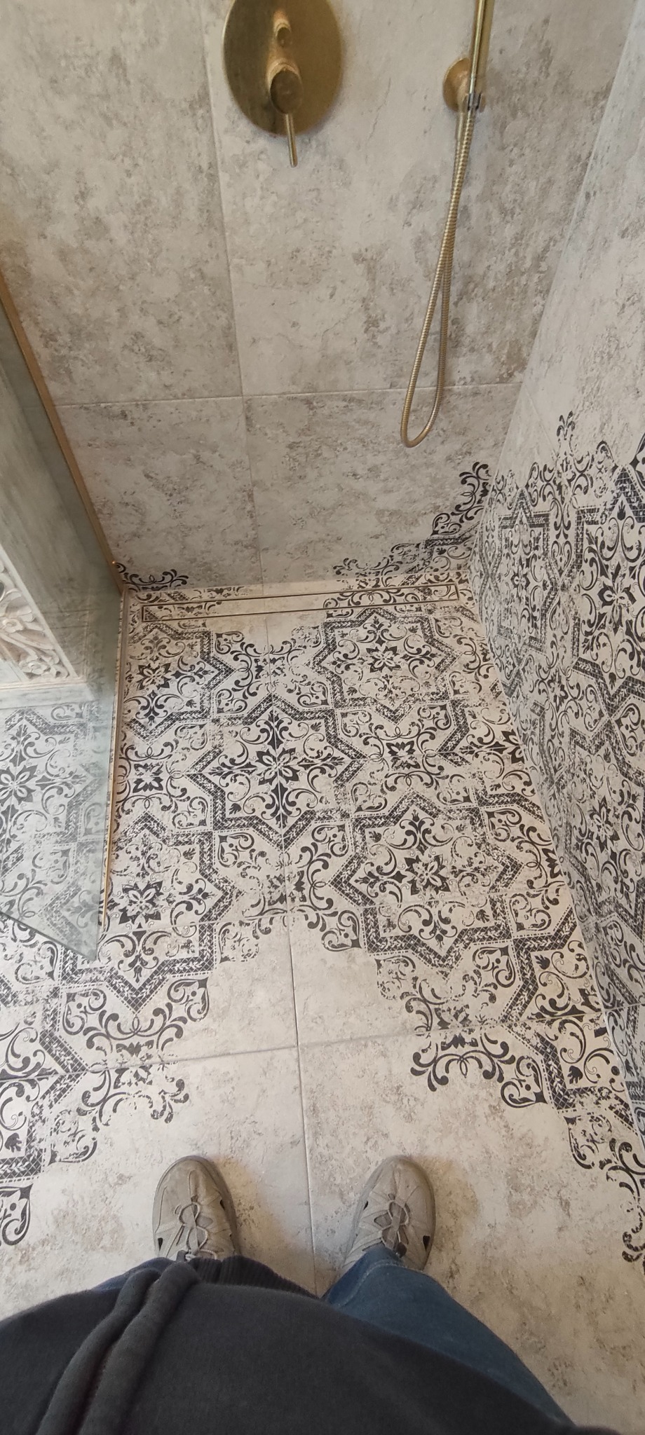 Wykończona kabina prysznicowa z dekoracyjnymi płytkami w odcieniach szarości i beżu, złota armatura. Widoczne stopy osoby robiącej zdjęcie.