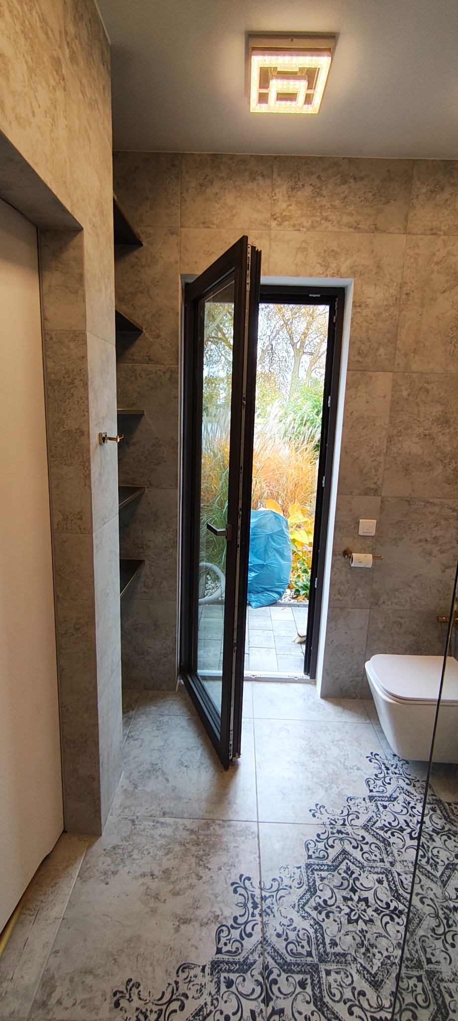 Wykończona łazienka z otwartymi drzwiami balkonowymi, przez które widać ogród. Na podłodze wzorzyste płytki, na ścianach płytki imitujące kamień. Widoczne półki i toaleta.