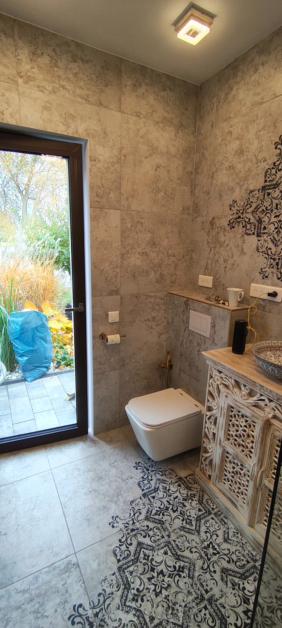 Wykończona łazienka z toaletą podwieszaną, płytkami imitującymi kamień i dekoracyjnym wzorem na podłodze. Widok na ogród przez przeszklone drzwi.