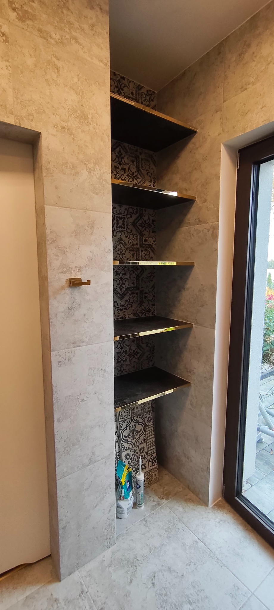 Narożna półka w łazience wyłożonej płytkami imitującymi kamień, z czarnym wnętrzem i złotymi krawędziami. Widoczne akcesoria łazienkowe i fragment okna.