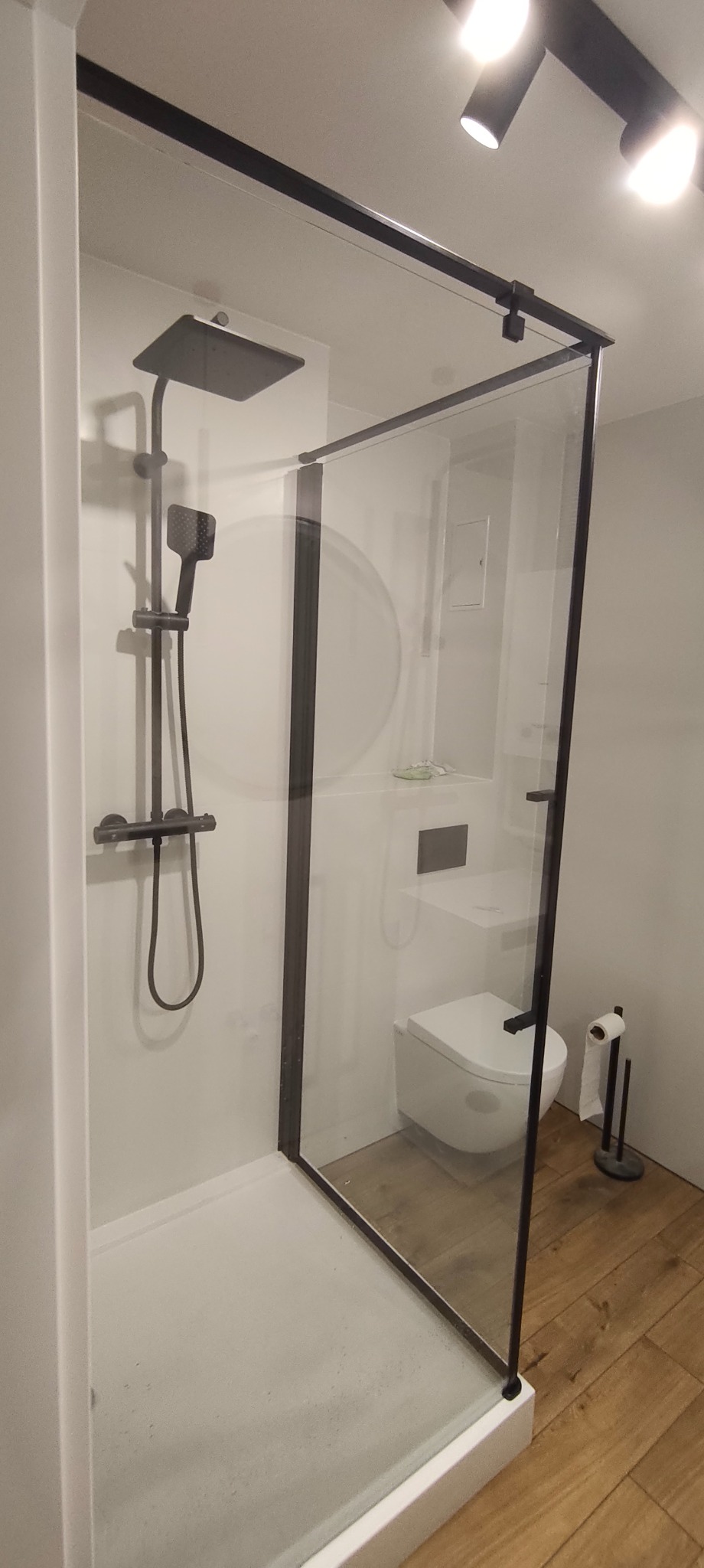 Nowoczesna kabina prysznicowa z czarnymi profilami i deszczownicą, widok na toaletę i drewnopodobną podłogę. Minimalistyczny design łazienki.