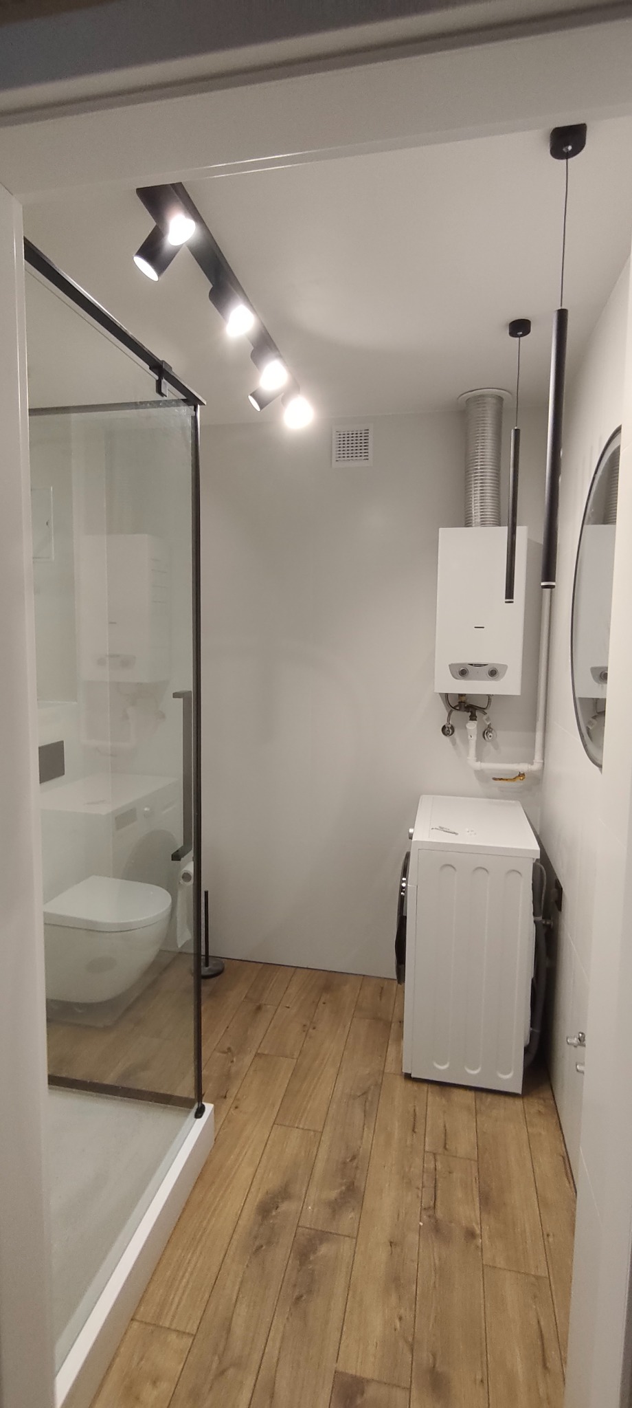 Nowoczesna łazienka z kabiną prysznicową typu walk-in, drewnopodobną podłogą, białymi ścianami i oświetleniem punktowym. Widoczny bojler i pralka.