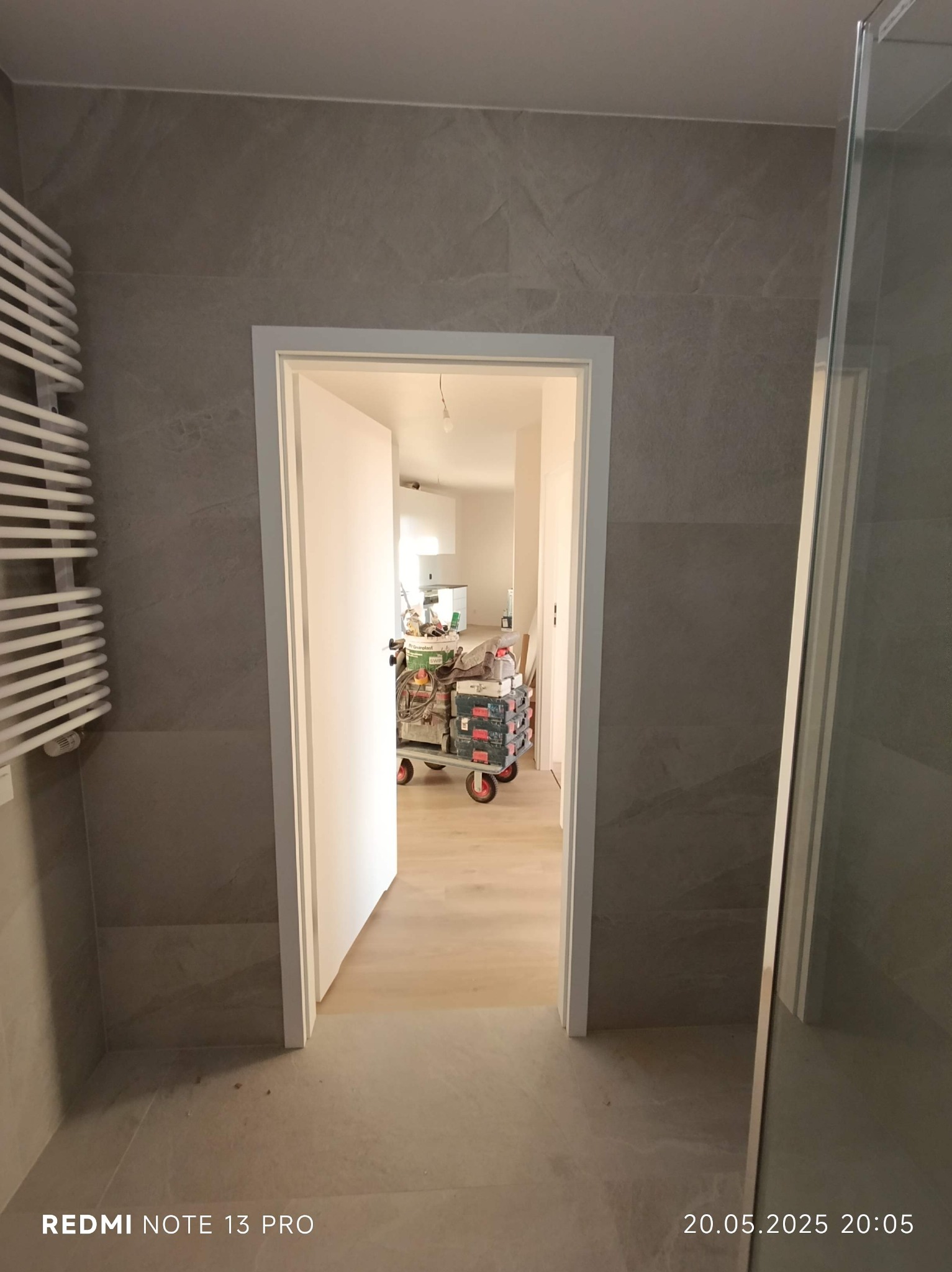 Wykończona łazienka z szarymi płytkami, widok przez otwarte drzwi na wózek z narzędziami. Grzejnik drabinkowy po lewej, kabina prysznicowa po prawej.