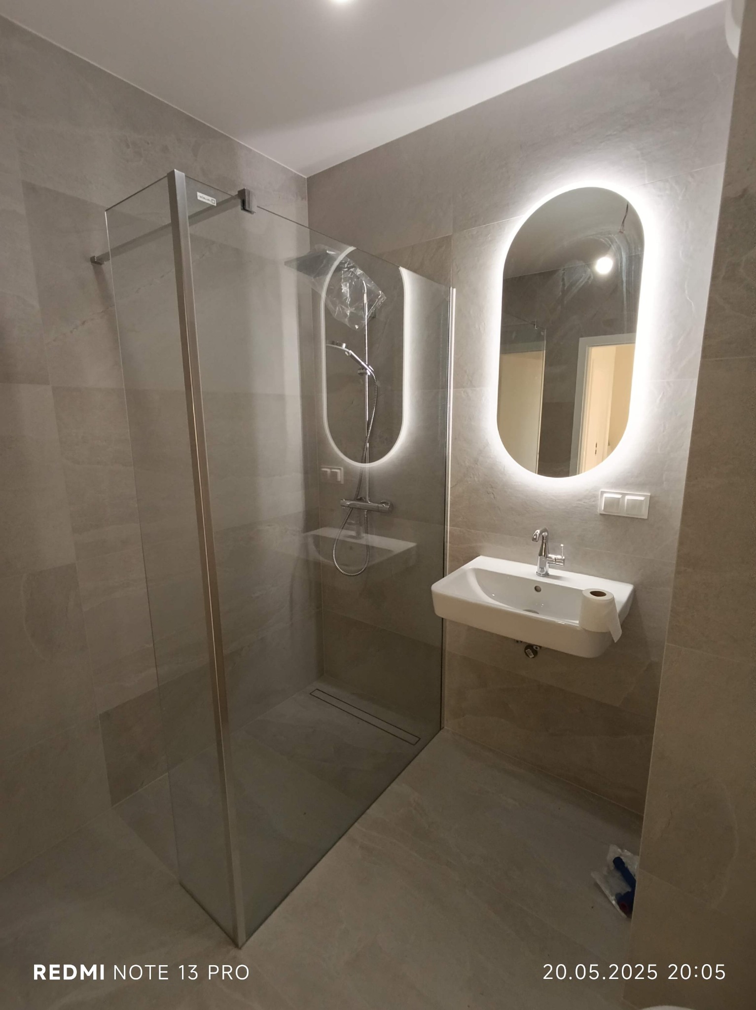 Nowoczesna łazienka w beżowych tonacjach: przeszklona kabina prysznicowa, umywalka z podświetlanym lustrem. Minimalistyczny design, stonowana kolorystyka, widoczny odpływ liniowy.