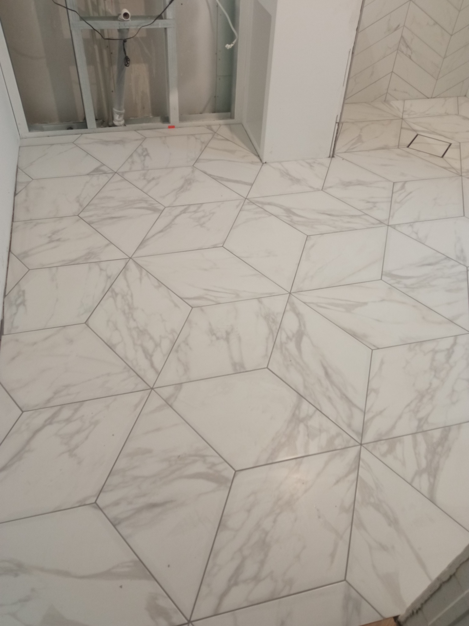 Ukończona podłoga w łazience z białych, heksagonalnych płytek imitujących marmur, ułożonych w geometryczny wzór 3D, widoczny odpływ liniowy i fragment ściany z płytek ułożonych w jodełkę.