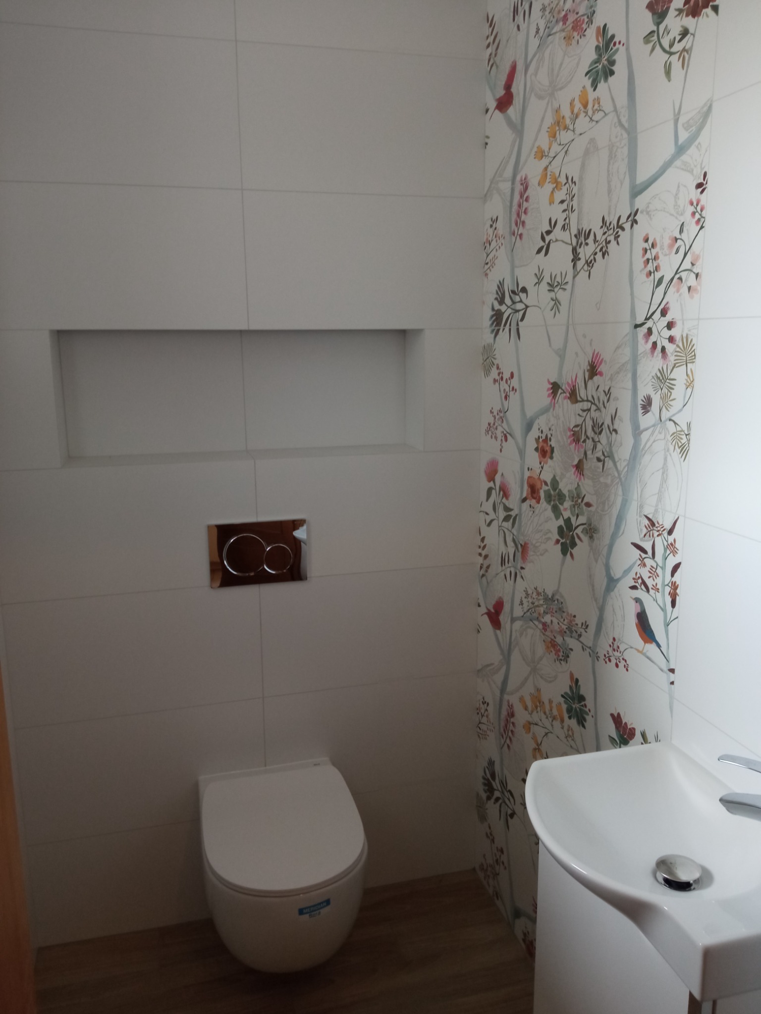 Nowoczesna łazienka z białymi płytkami, wiszącą toaletą, umywalką i akcentem w postaci tapety w kwiaty i ptaki na jednej ze ścian.