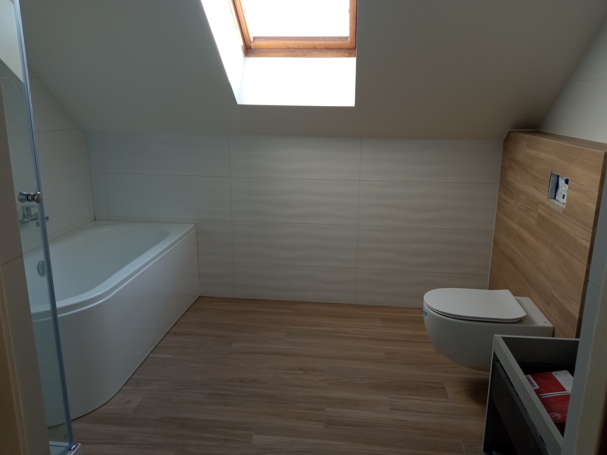 Nowoczesna łazienka na poddaszu z białą wanną, wiszącą toaletą z deską wolnoopadającą, drewnopodobną podłogą i oknem dachowym.