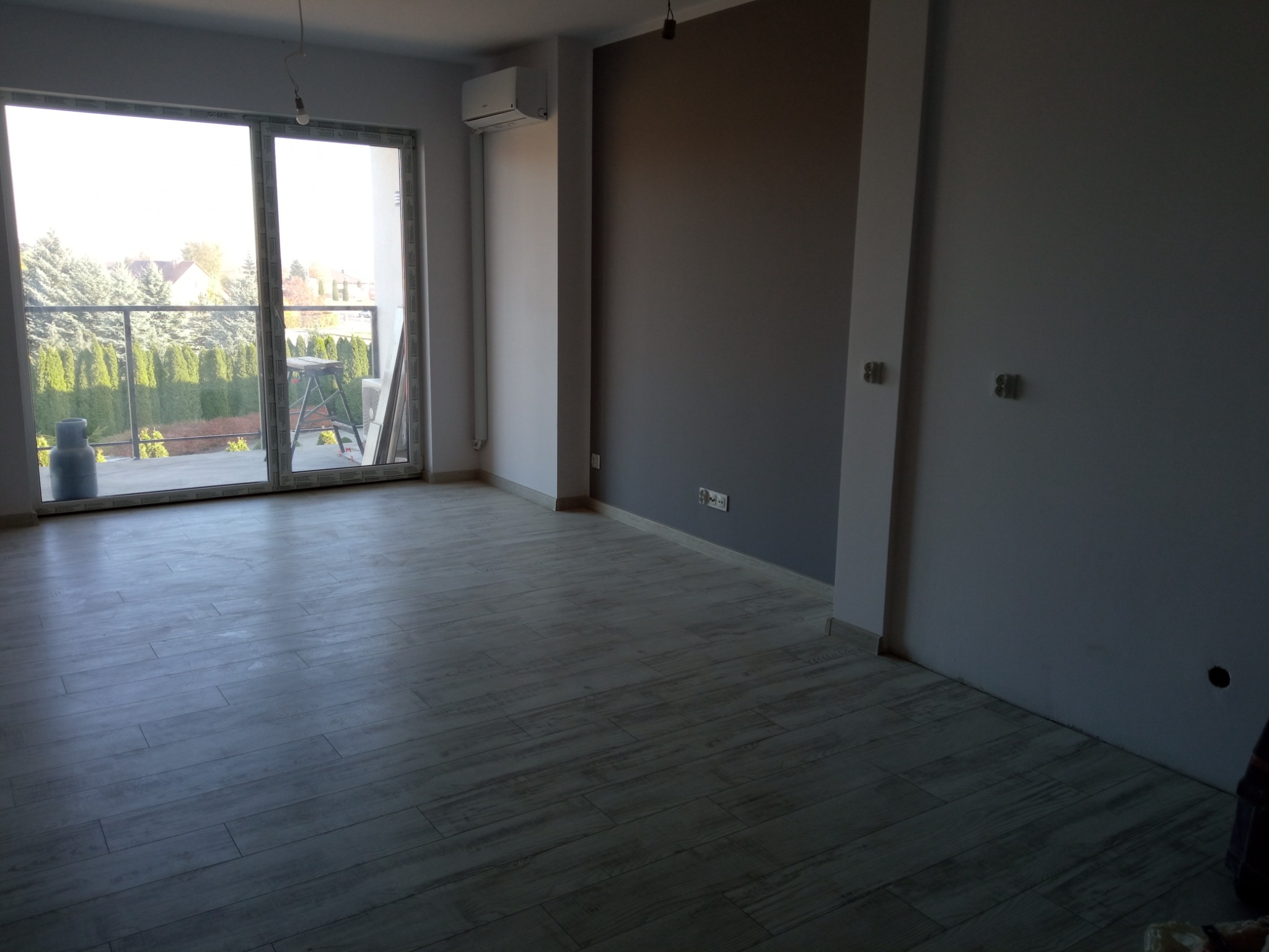 Pomalowane na szaro ściany i ułożona podłoga z paneli imitujących drewno w pustym pokoju z dużym oknem balkonowym w Wolicy.