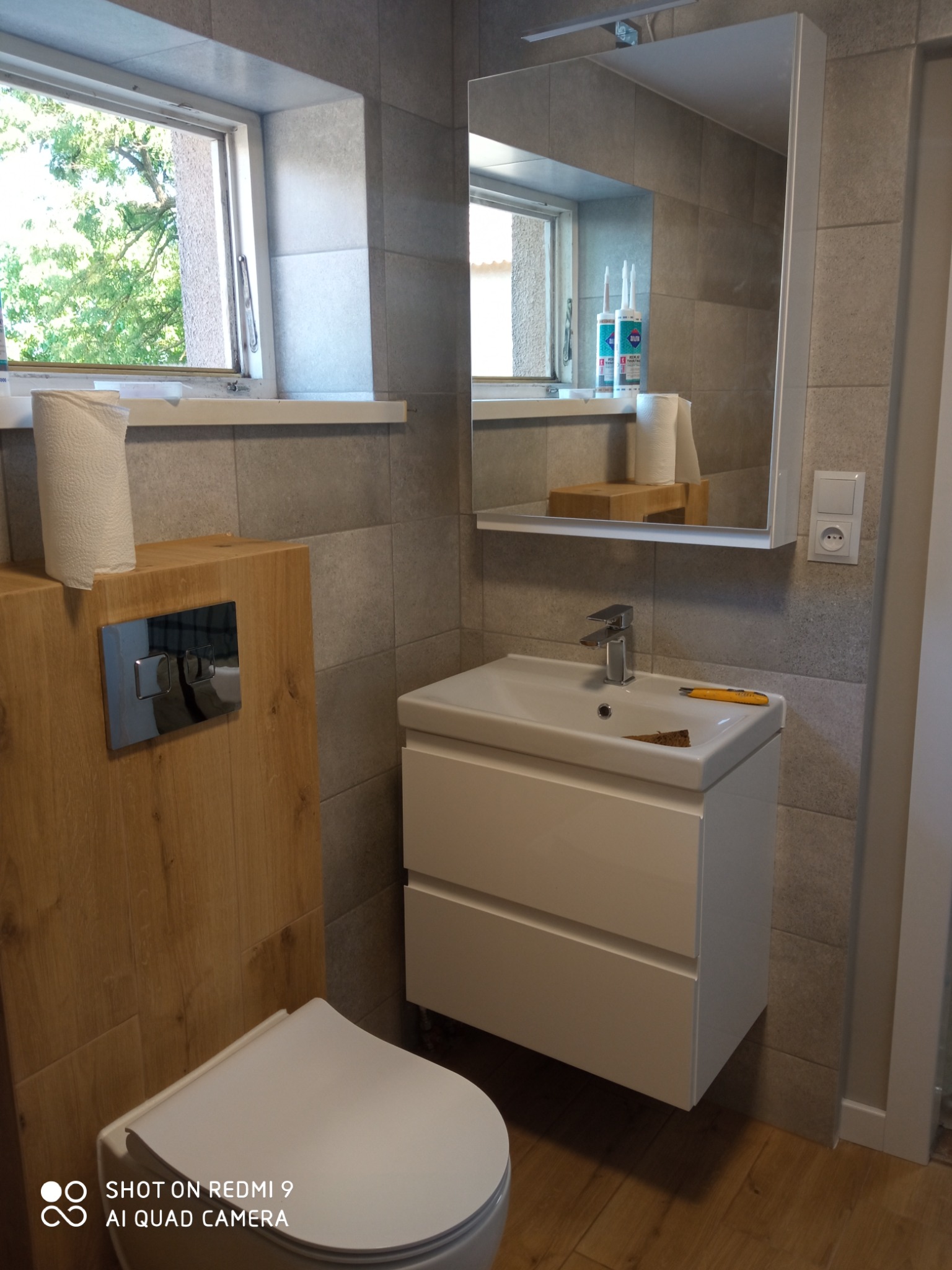 Nowoczesna łazienka z wiszącą szafką pod umywalką, drewnianą obudową WC, szarą glazurą i lustrem z szafką, widoczne narzędzie na umywalce.