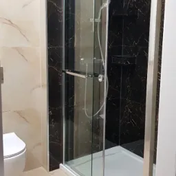 Nowoczesna kabina prysznicowa z przesuwnymi drzwiami, chromowanymi detalami i czarnymi płytkami imitującymi marmur, uzupełniona beżowymi płytkami podłogowymi i wiszącą toaletą.