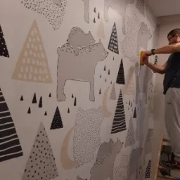 Mężczyzna na drabinie wygładza żółtą packą tapetę z motywem niedźwiedzi i geometrycznych wzorów na ścianie.