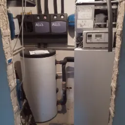 Zestaw urządzeń grzewczych Altech z zbiornikiem na wodę, widoczne czarne rury, szara obudowa, manometry oraz fragment drabiny w tle, instalacja w trakcie prac wykończeniowych.