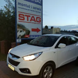 Biały Hyundai Tucson zaparkowany przed szyldem reklamowym firmy oferującej montaż i serwis samochodowych instalacji gazowych, widoczny znak STAG.