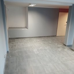 UR Dobra Ekipa - Pomalowane na szaro wnętrze biura z szarą podłogą z płytek i wnęką w ścianie, widoczne kable wystające ze ściany.