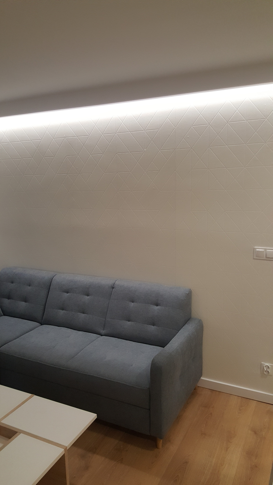 Szary narożnik w salonie z drewnianą podłogą, białą ścianą z geometrycznym wzorem i podświetleniem LED pod sufitem.