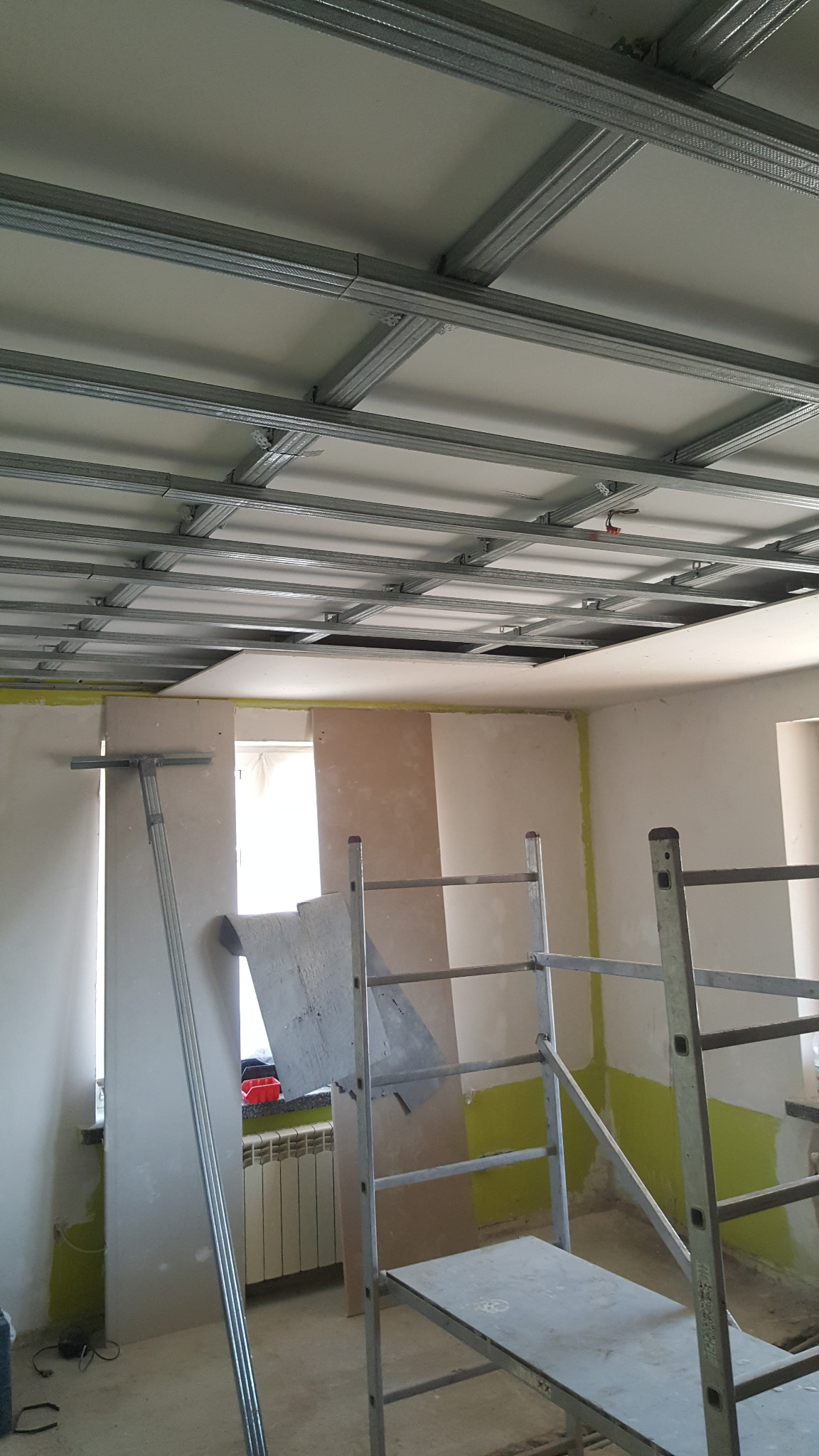 Montaż metalowej konstrukcji sufitu podwieszanego w pomieszczeniu w trakcie prac, widoczne płyty gipsowo-kartonowe, drabina i narzędzia.
