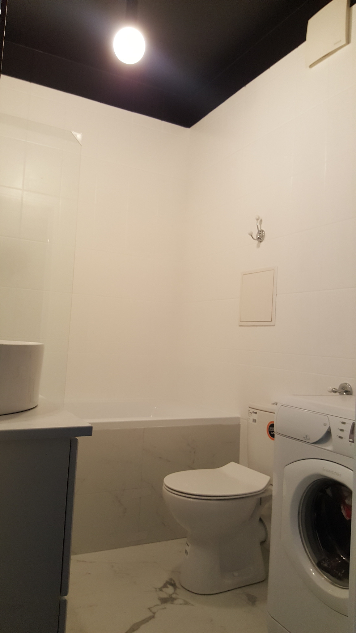 Jasna łazienka z wanną, umywalką, toaletą i pralką, wykończona białymi płytkami z szarymi żyłkami na podłodze i ścianach, z czarnym sufitem i punktowym oświetleniem.