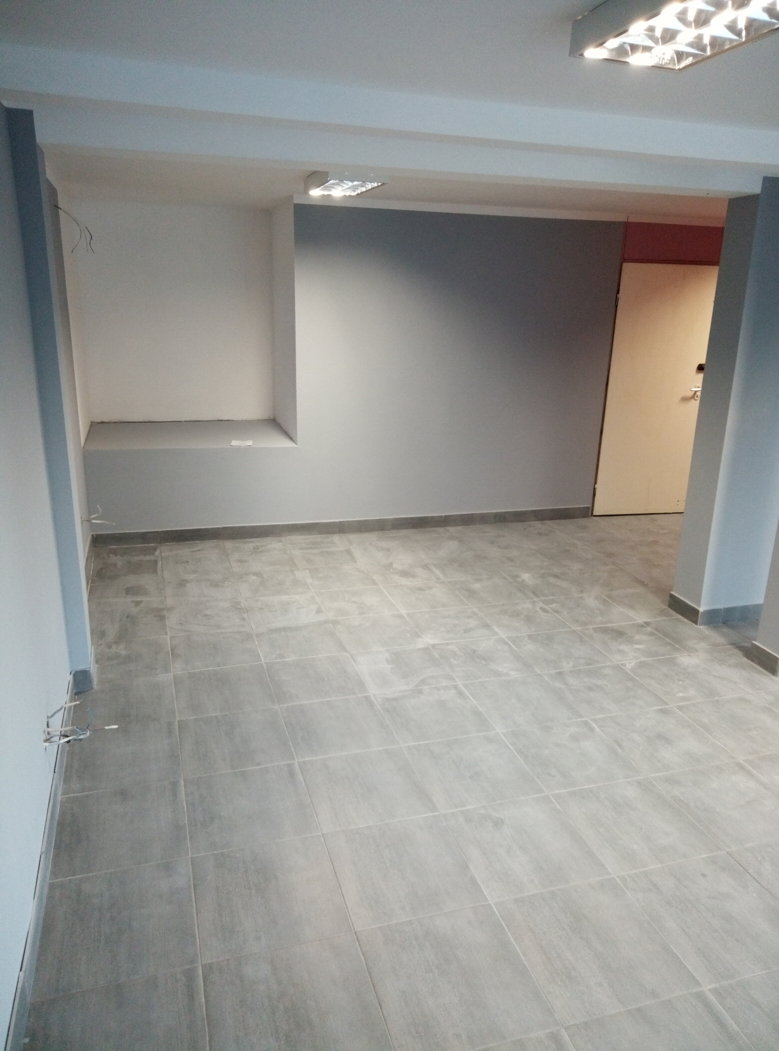 Pomalowane na szaro wnętrze biura z szarą podłogą z płytek i wnęką w ścianie, widoczne kable wystające ze ściany.