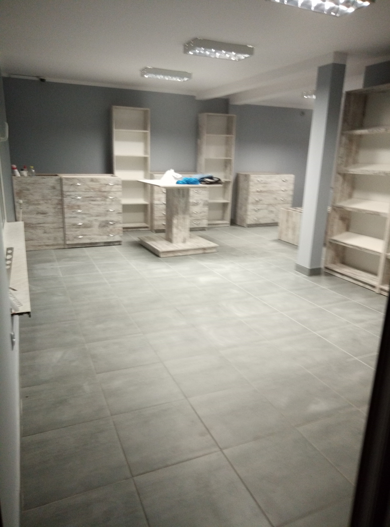 Jasne biuro z szarymi ścianami, podłogą z płytek i drewnianymi regałami oraz komodami w stylu vintage, częściowo umeblowane, z niebieskim materiałem leżącym na stole.