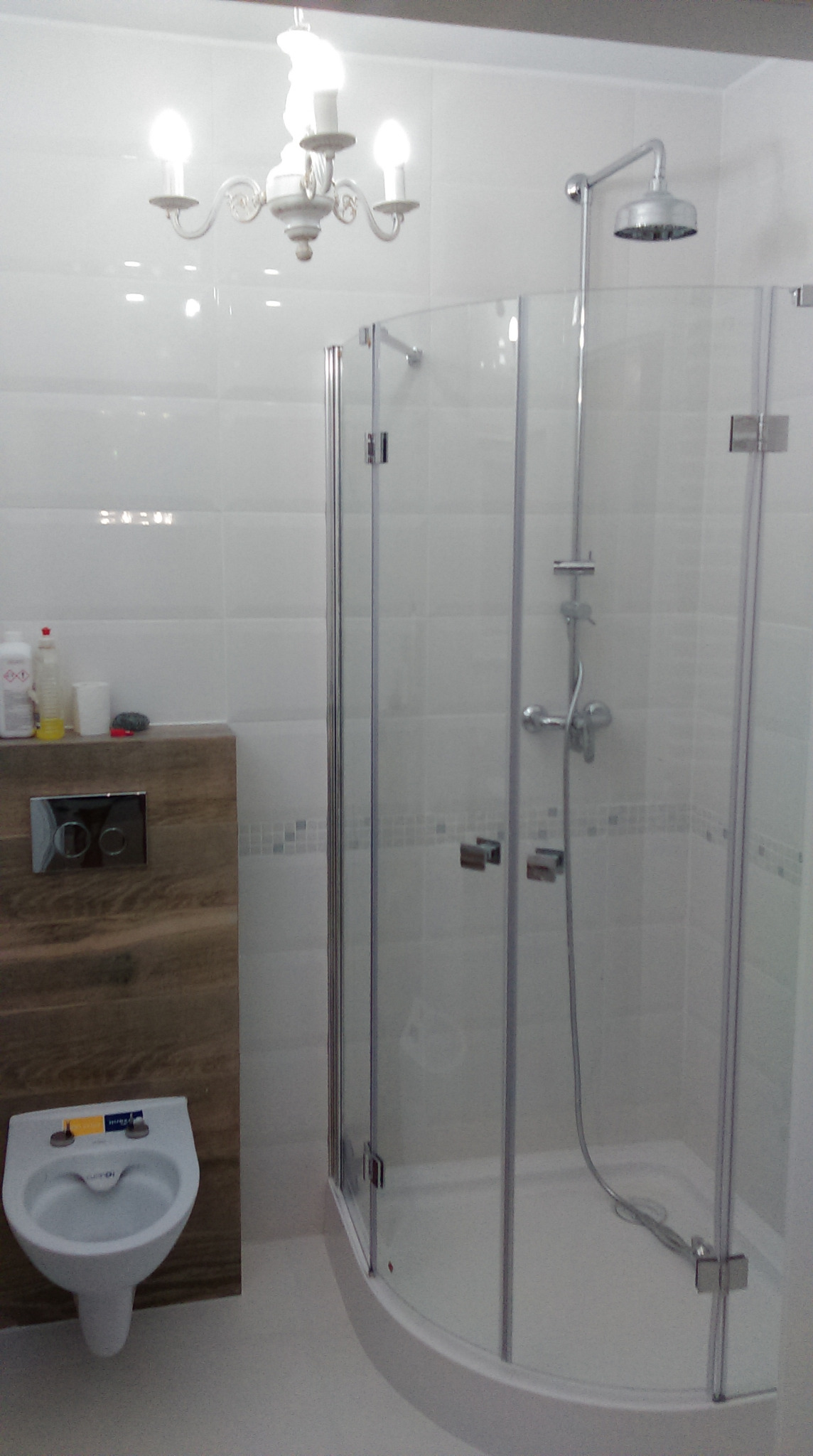 Nowoczesna łazienka z kabiną prysznicową z transparentnego szkła, białymi płytkami, podwieszaną muszlą klozetową i ozdobnym żyrandolem.