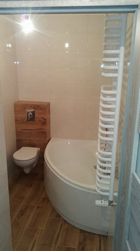 Nowoczesna łazienka po remoncie z narożną wanną, drewnopodobną zabudową toalety i grzejnikiem drabinkowym.