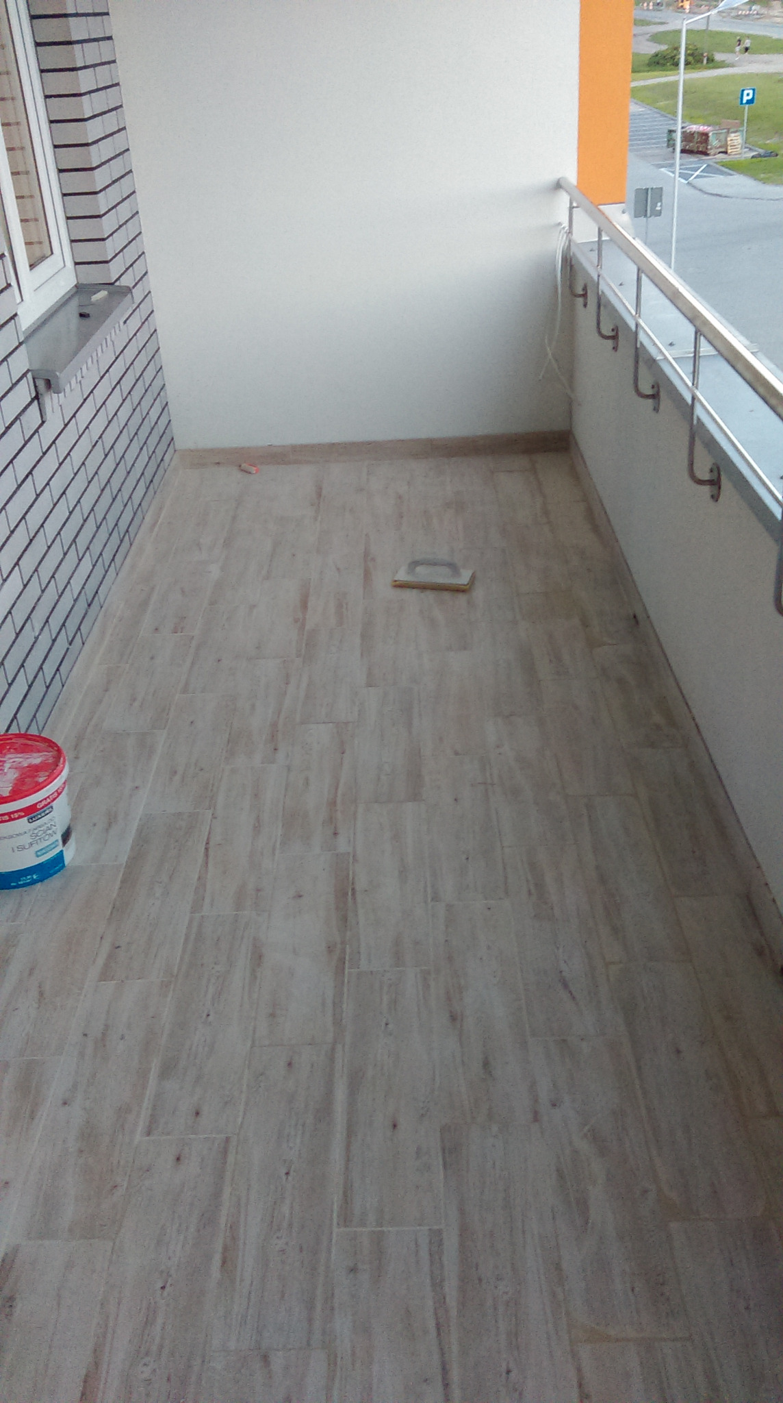 Balkon z nowo ułożoną podłogą z płytek imitujących drewno, widoczne narzędzie do rozprowadzania kleju i wiadro z preparatem gruntującym, ściany pomalowane na biało.