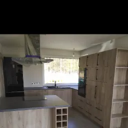 Wnętrze kuchni w trakcie montażu, z wyspą i drewnopodobnymi frontami, okap zabezpieczony folią, widoczne gniazdka elektryczne i duży czarny piekarnik.