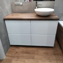 Biała szafka łazienkowa z czterema szufladami i drewnianym blatem, na którym stoi okrągła umywalka. W tle szare płytki i fragment wanny.