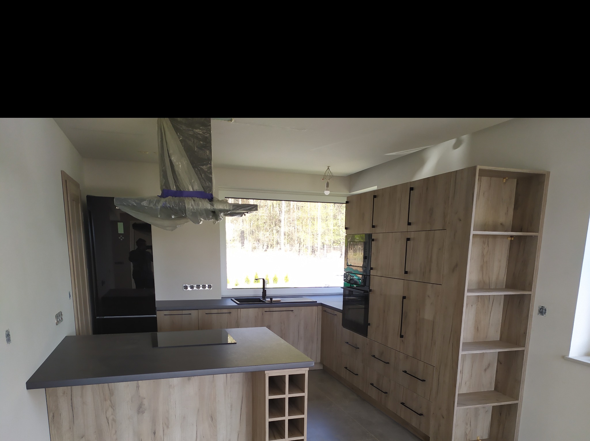 Wnętrze kuchni w trakcie montażu, z wyspą i drewnopodobnymi frontami, okap zabezpieczony folią, widoczne gniazdka elektryczne i duży czarny piekarnik.