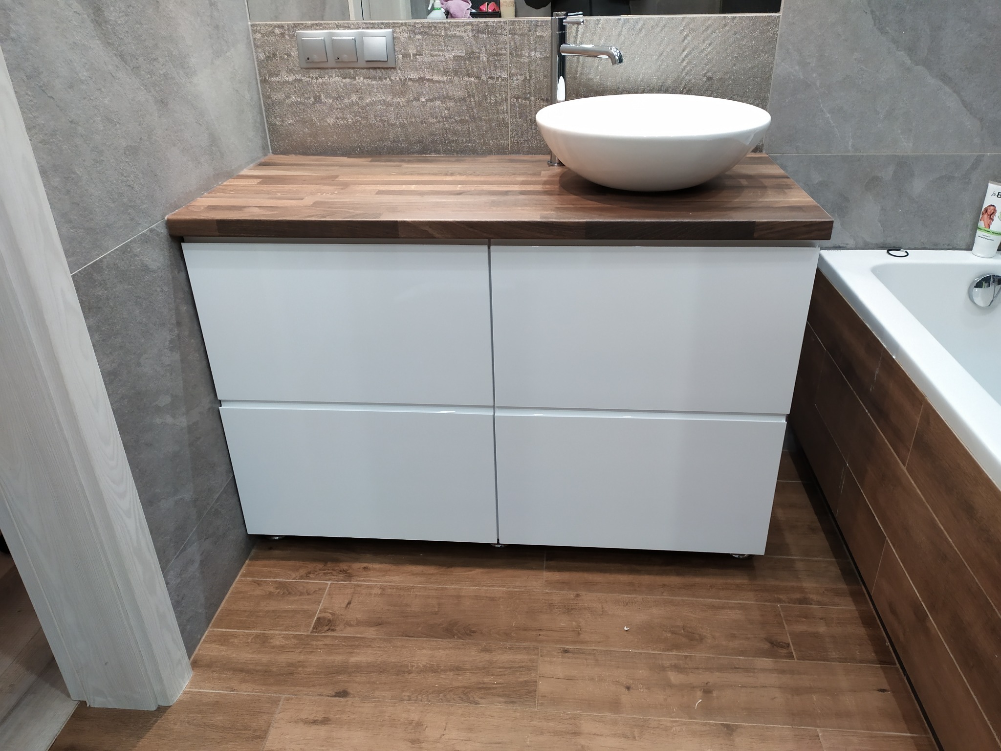 Biała szafka łazienkowa z czterema szufladami i drewnianym blatem, na którym stoi okrągła umywalka. W tle szare płytki i fragment wanny.