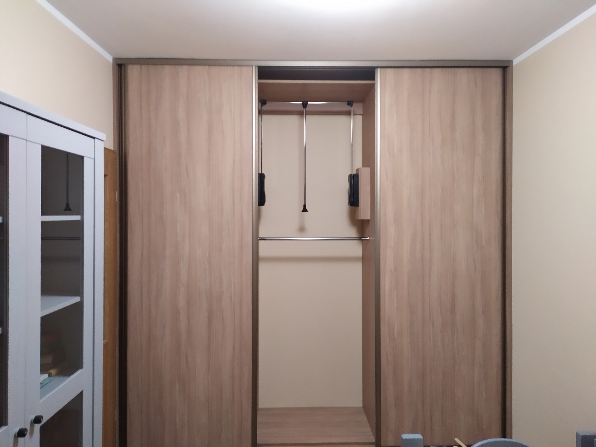 Wnętrze szafy przesuwnej z drążkiem i wysuwanymi wieszakami, widoczna z boku biała witryna z półkami.