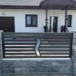 Nowoczesne, poziome przęsło ogrodzeniowe z metalu w kolorze grafitowym, zamontowane na murku z szarych bloczków betonowych, na tle białego domu z ciemnymi elementami wykończenia.