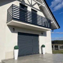 Nowoczesny dom z szarą, poziomą balustradą balkonową, szarą bramą garażową rolowaną i dwoma białymi donicami z kulistymi krzewami.