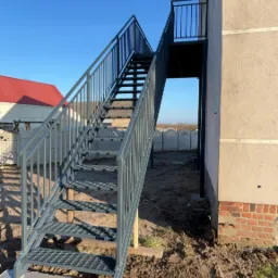 Zewnętrzne schody metalowe z ażurowymi stopniami, prowadzące na balkon, pomalowane na szaro, osadzone na betonowej podstawie, z widokiem na dom z czerwoną dachówką i błękitne niebo.